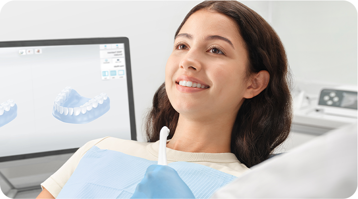 Patient in a dental chair as a clinician holds an intraoral scanner, with a 3D teeth model on a monitor