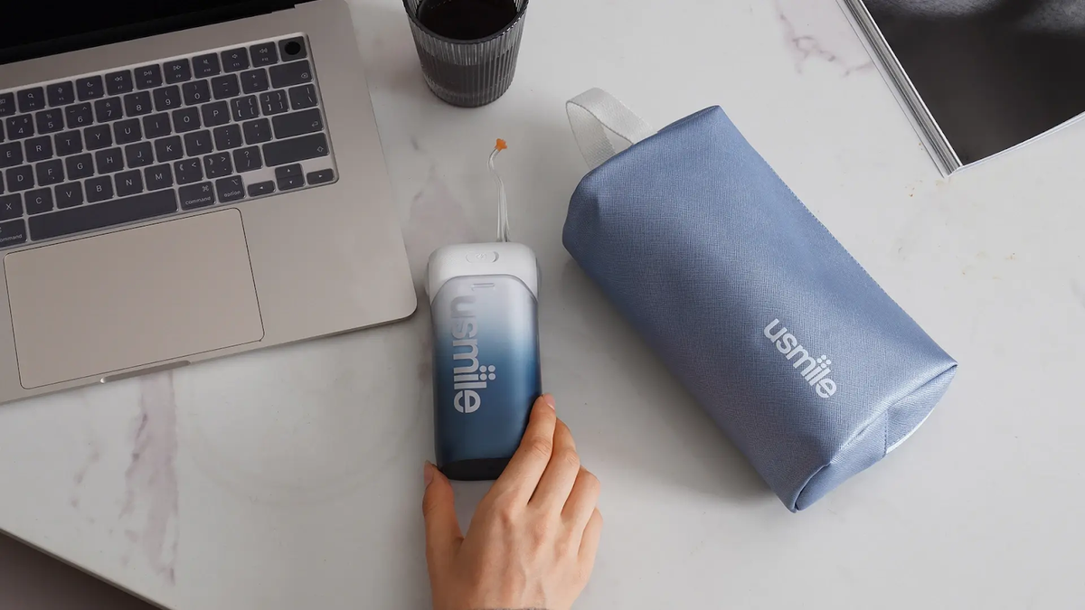 Hand holding a compact blue usmile water flosser beside its travel pouch and a laptop